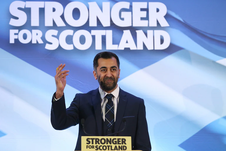 Hamza Yousaf waving in front of a sign that reads 'stronger for Scotland'