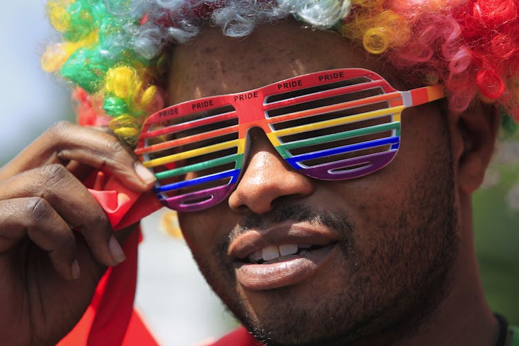 A black man in a rainbow wig with rainbow sunglasses.