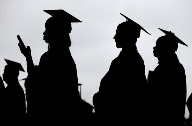 Silhouetted students in caps and gowns seen at a graduation, one holds a smart phone