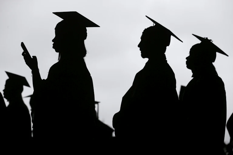 Silhouetted students in caps and gowns seen at a graduation, one holds a smart phone