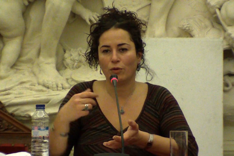 View of a brunette woman talking in front of a mic against the backdrop of a sculpted mural