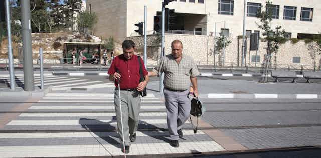 Man helps blind man with cane to cross the street.