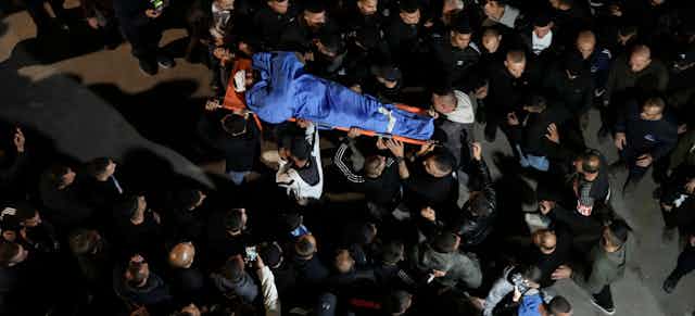 A crowd of people carry the body of a man wrapped in a blue shroud.