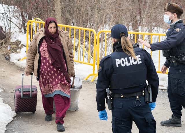 A woman pulling a suitcase and in a hijab is stopped by police on a snowy road.