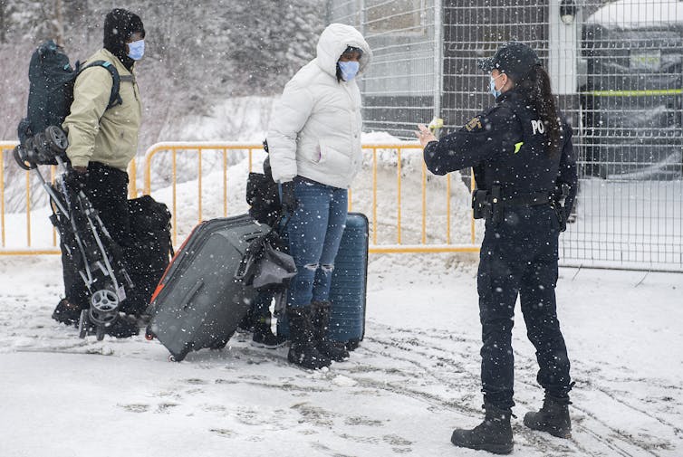 A police officer with a ponytail stops people bundled in parkas and pulling suitcases along a snowy road.