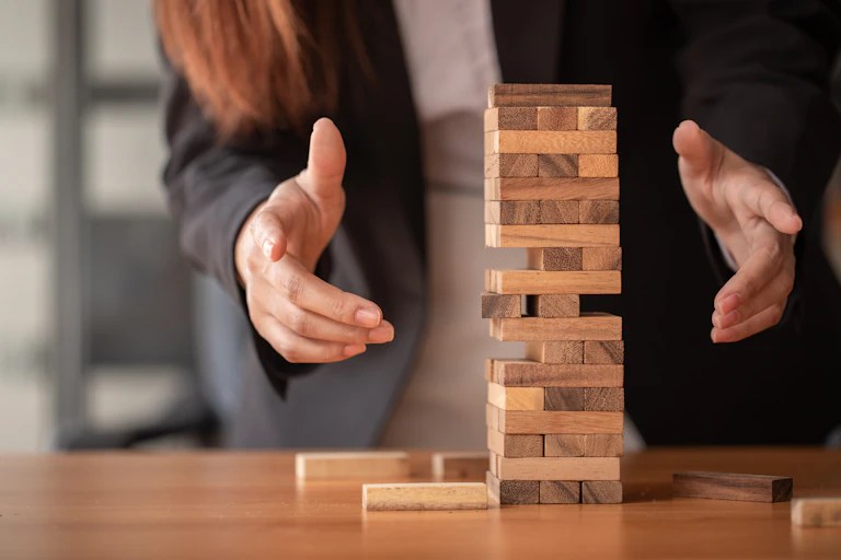 Jenga tower with missing pieces, still standing, two hands around the tower, stopping it from collapsing.