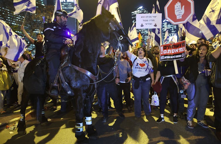 A crowd of people carryg signs and flags and shout as a police officer on a horse stands in front of them during the night.
