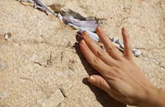 A woman's hand delicately pushes a written note into a crack in a stone wall.