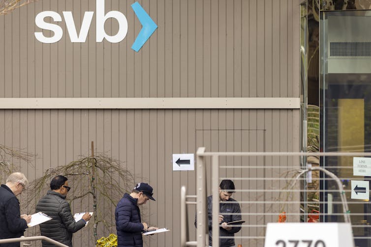 People line up, social distanced, along a wall with the letters s, v and b.