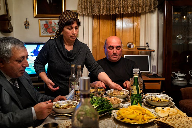 A woman in a headscarf puts plates full of soup on a table in front of two men.