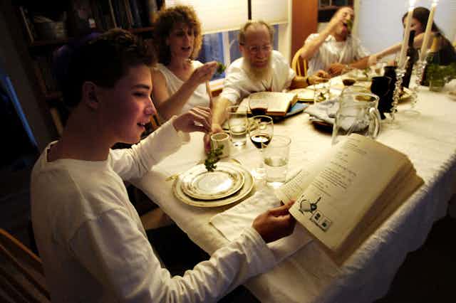 A young man in a white shirt reads from a book as he holds a sprig of parsley at a family meal.