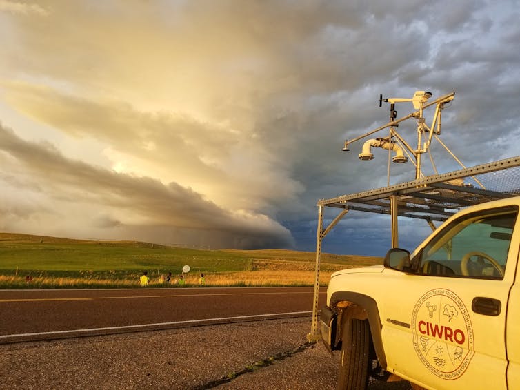 Scientists stand near a truck outfitted with measuring devices with a dramatic storm on the horizon.