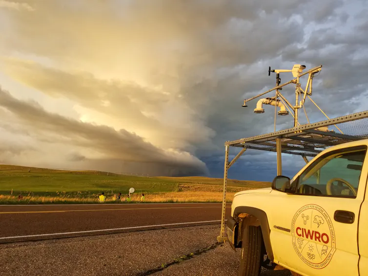 Scientists stand near a truck outfitted with measuring devices with a dramatic storm on the horizon.