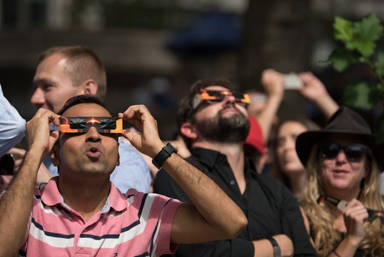 Several people wearing black paper glasses looking at the sky in awe