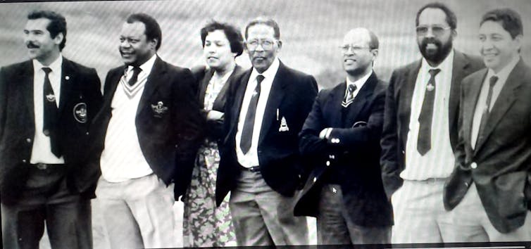 A black and white photograph of a row of six men and a woman standing posing, the men wearing jackets and ties