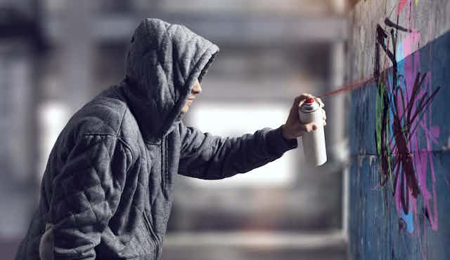 A young person in a hooded sweatshirt sprays graffiti on a cement wall
