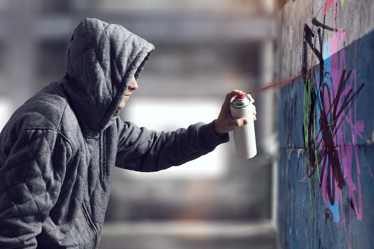 A young person in a hooded sweatshirt sprays graffiti on a cement wall