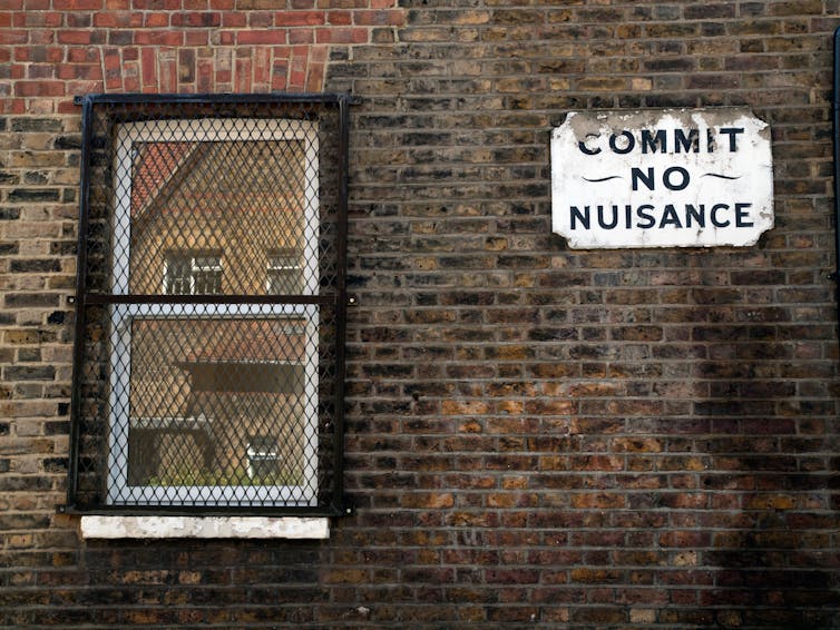 An exterior brick wall with an aged sign reading Commit No Nuisance.