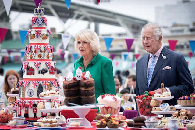 Charles and Camilla stand at a table filled with strawberries, baked treats, and a seven-tier cake decorated with symbols of London and the queen.