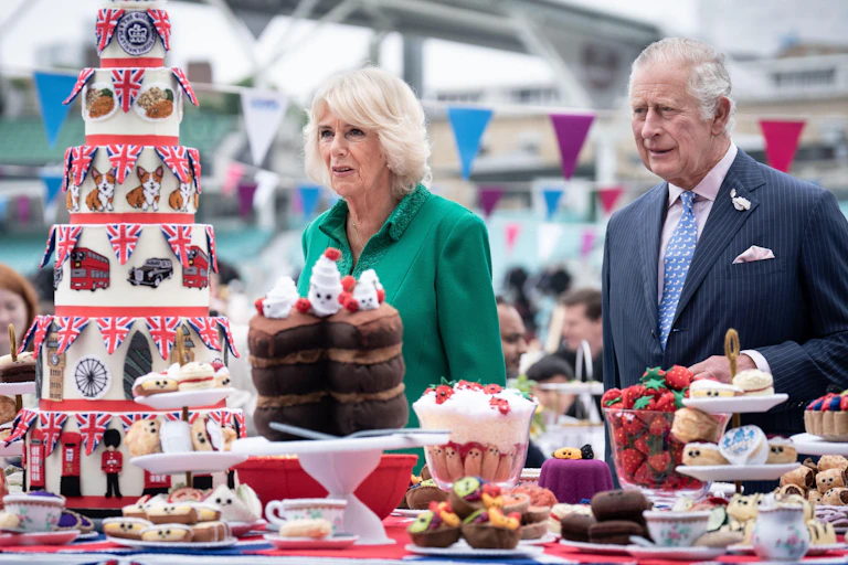 Charles and Camilla stand at a table filled with strawberries, baked treats, and a seven-tier cake decorated with symbols of London and the queen.