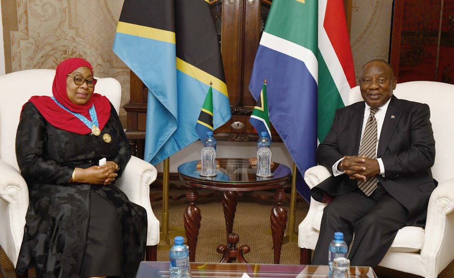 A man and a woman cusp their hands while sitting. Behind them are the flags of Tanzania and South Africa.