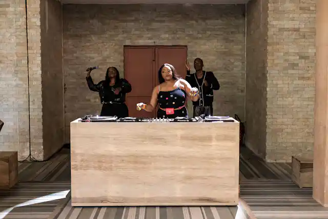An African woman in a sleeveless black dress with logos on performs behind deejay decks. Behind her, a man and a woman dance, holding microphones.
