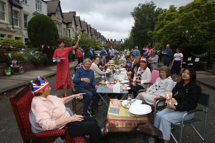 People, some wearing paper crowns or waving flags, gather around a long picnic table in a street.