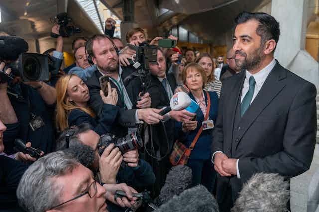 A smiling Humza Yousaf surrounded by journalists pointing microphones. 