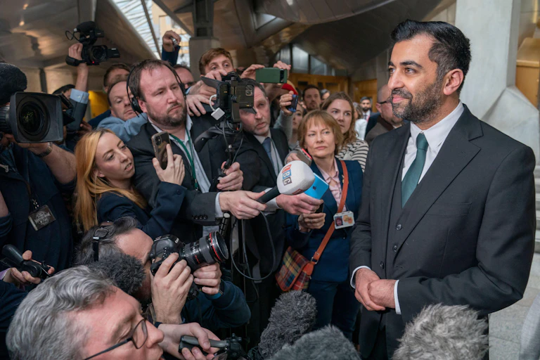 A smiling Humza Yousaf surrounded by journalists pointing microphones.