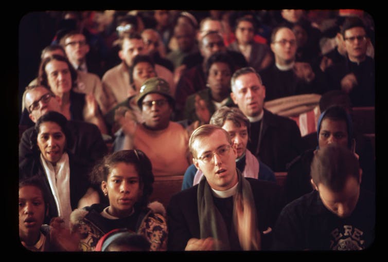A photograph shows a crowd sitting in church pews as a man in a clerical scholar speaks.