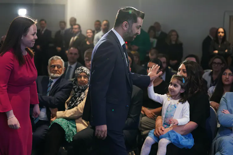 Humza Yousaf high fiving his daughter as he walks past. She is sitting on her mother's knee in a crowded room.