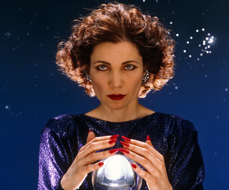 Mystic Meg's hair is blow backwards as she wraps her hands (with long red fingernails) around a crystal ball. 