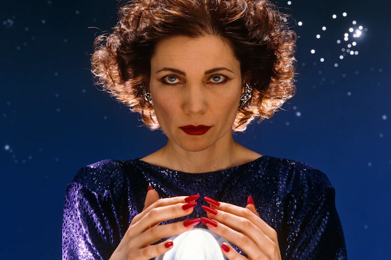 Mystic Meg's hair is blow backwards as she wraps her hands (with long red fingernails) around a crystal ball.