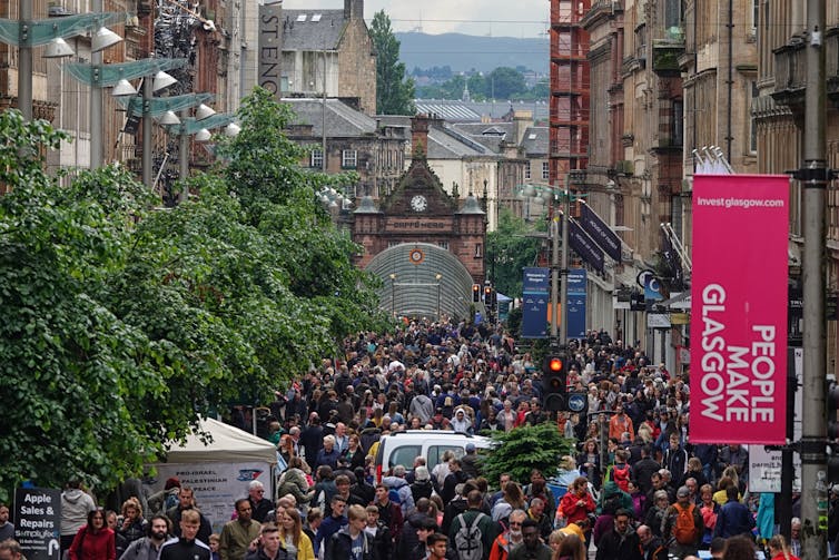 The famous shopping district in the city, Buchanan Street, is shown filled with people during an afternoon day. A pink sign on a lamp post says 