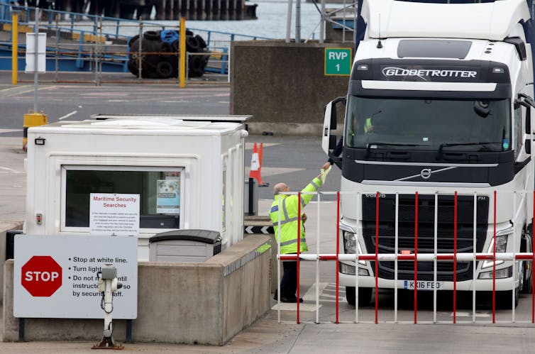 A large commercial truck stops at a gate with a booth and a person holding a piece of paper.