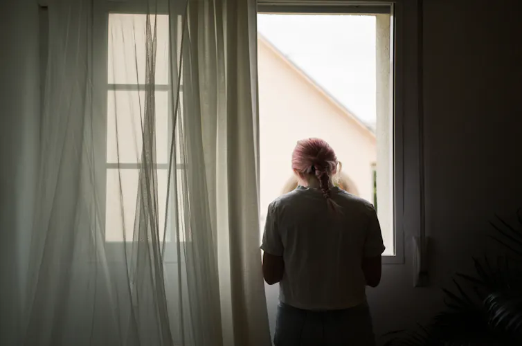 A young woman facing away from the camera looks out of a window.
