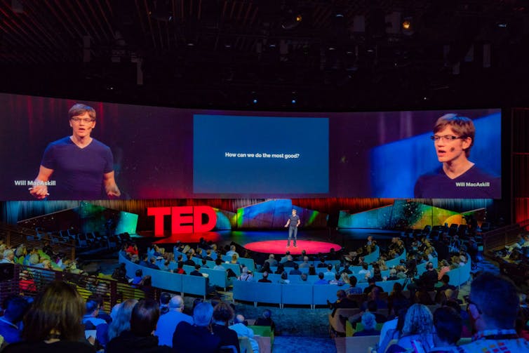 A man in a dark-colored t-shirt speaks on a stage in front of a live audience, with two massive screens behind him.