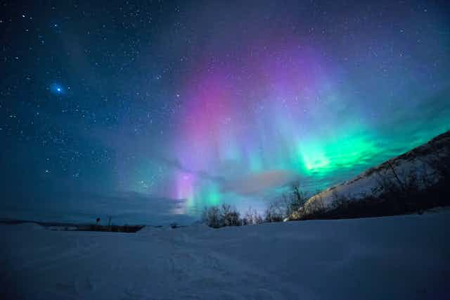 A photo of green, blue and purple aurora against a starry sky over a snow-covered landscape.