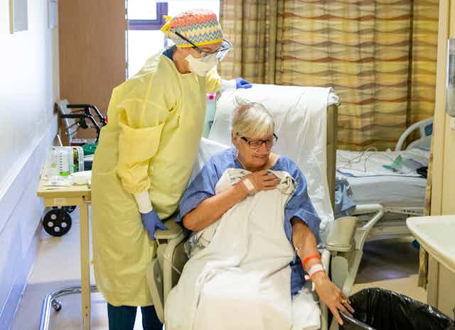 A personal care worker in yellow PPE helps a woman in a blue hospital gown