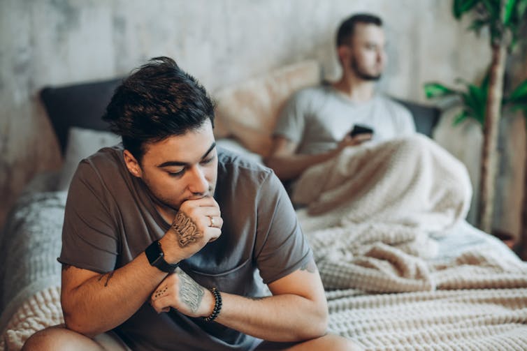Male couple, one sitting in bed, the other sitting on edge of bed, looking worried