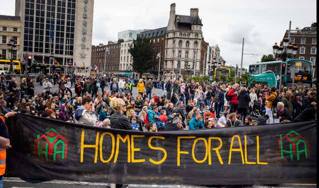 People sitting on a Dublin street behind a large black banner that reads "Homes for all" in yellow letters.