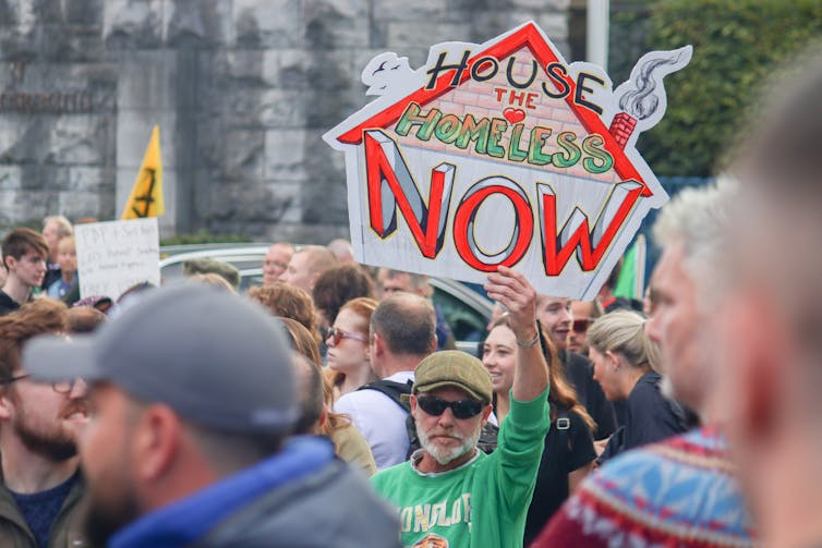 A man in a green top, hat and sunglasses, in a crowd of people, holds up a sign reading ‘House the homeless now’.