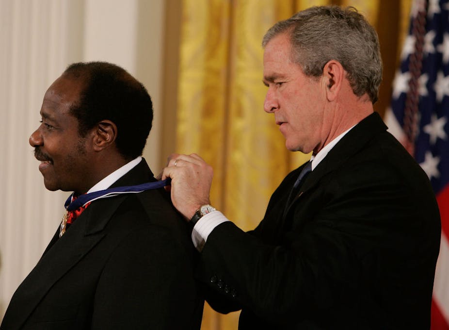 A man tying a ribbon around another man's neck, with the American flag behind them 