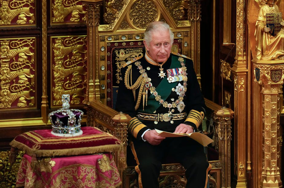 King Charles sits in a gilded golden throne next to the crown. He wears formal military regalia. 