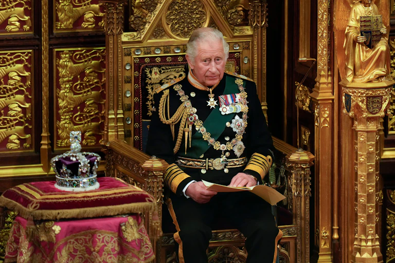 King Charles sits in a gilded golden throne next to the crown. He wears formal military regalia.