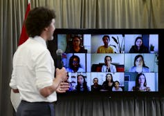 A dark-haired man in a white shirt faces a video screen of people, including a woman with children.