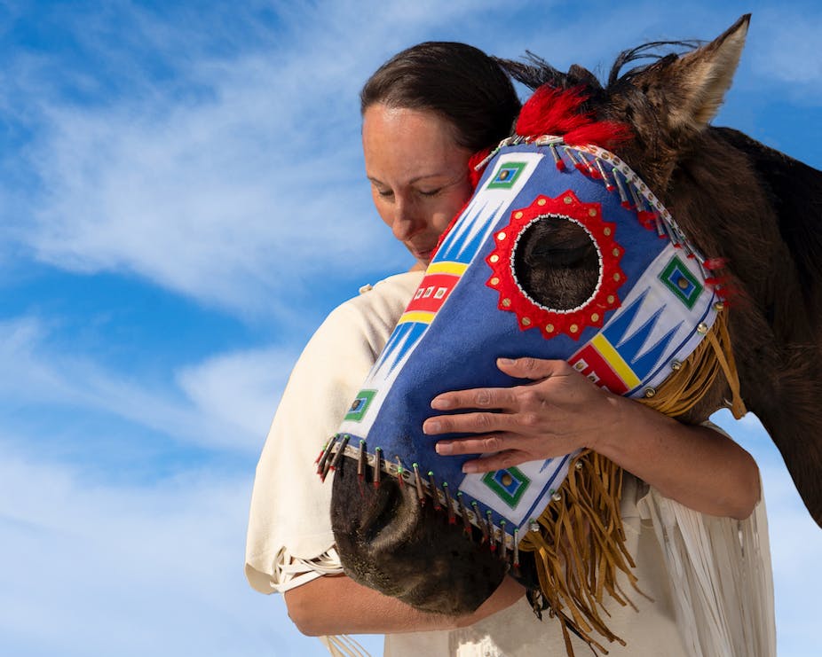 Lakota woman leans on horse wearing decorative mask