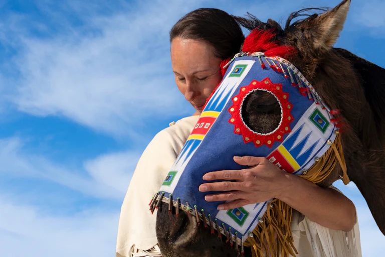 Lakota woman leans on horse wearing decorative mask