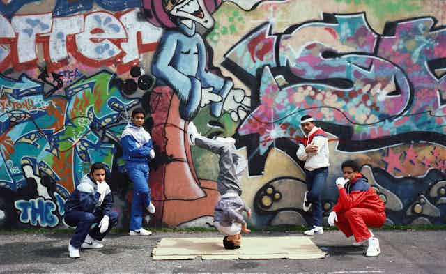 A group of five men stand in front of a wall covered in grafitti as one of them spins on his head.