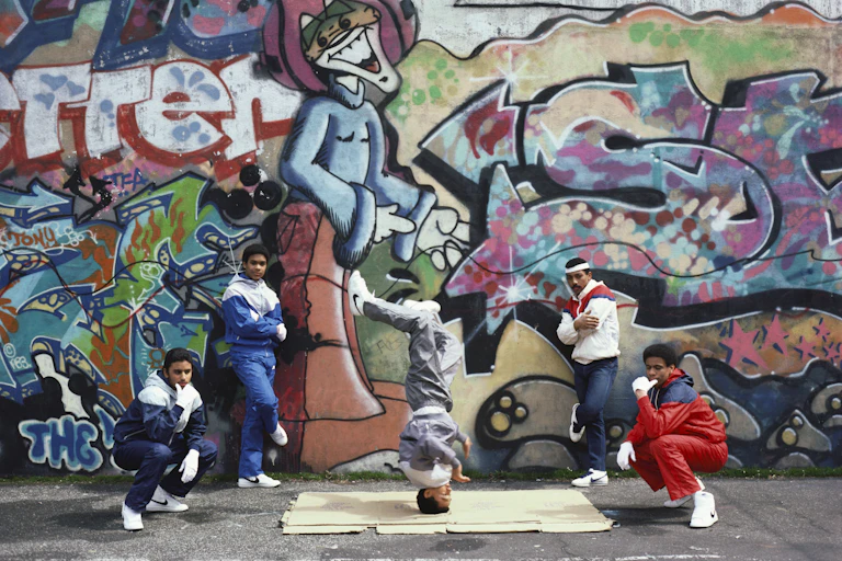 A group of five men stand in front of a wall covered in grafitti as one of them spins on his head.